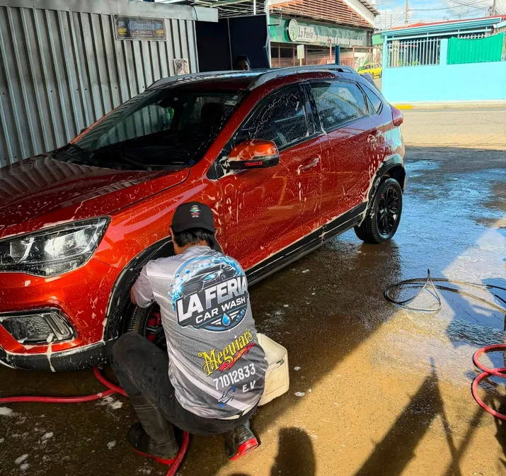 Un trabajador con camisa de La Feria Car Wash lava a mano las llantas de un carro rojo cubierto de espuma